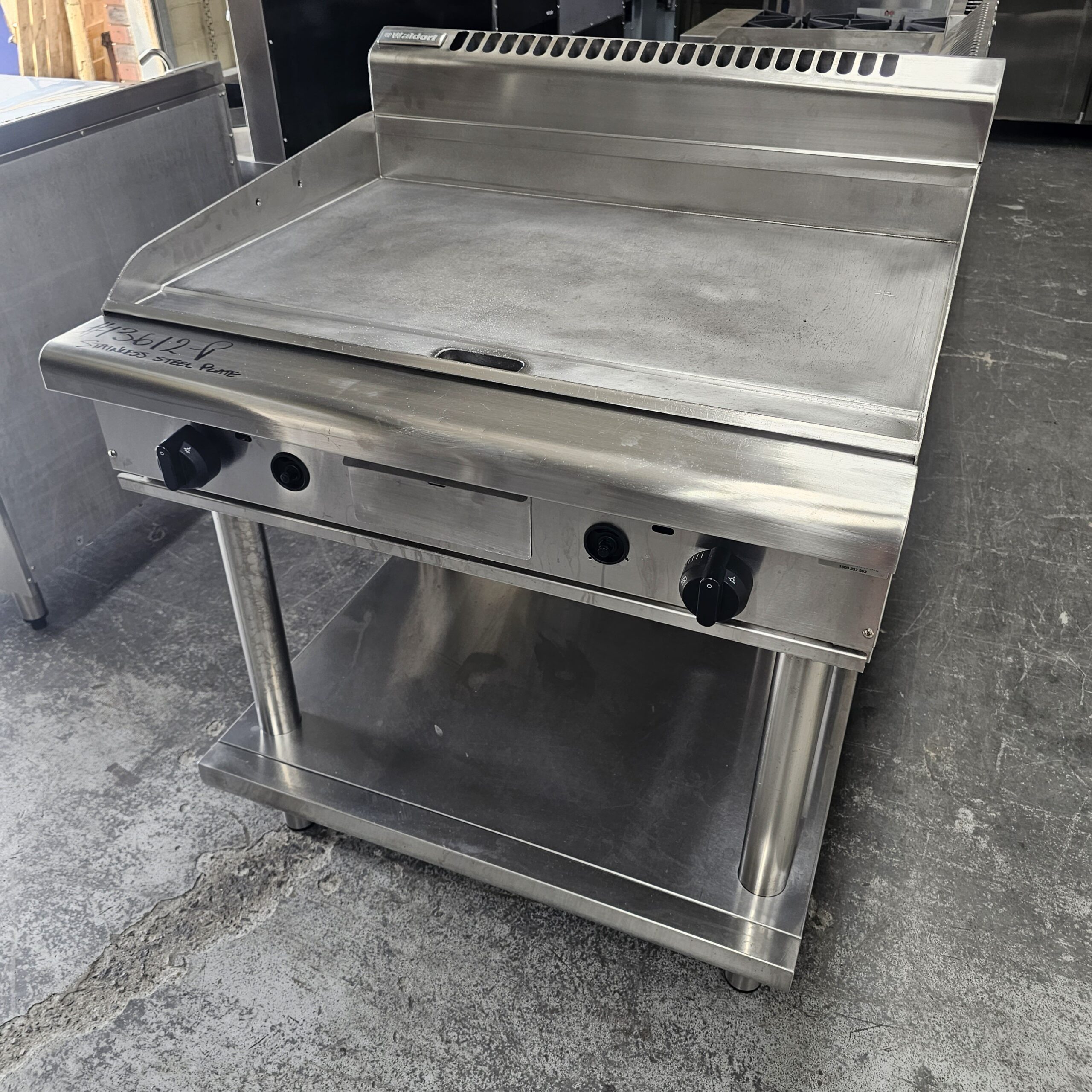 A commercial stainless steel flat-top griddle with two control knobs, a backsplash, and an open shelf underneath, placed on a concrete floor in an industrial kitchen setting. MRCE