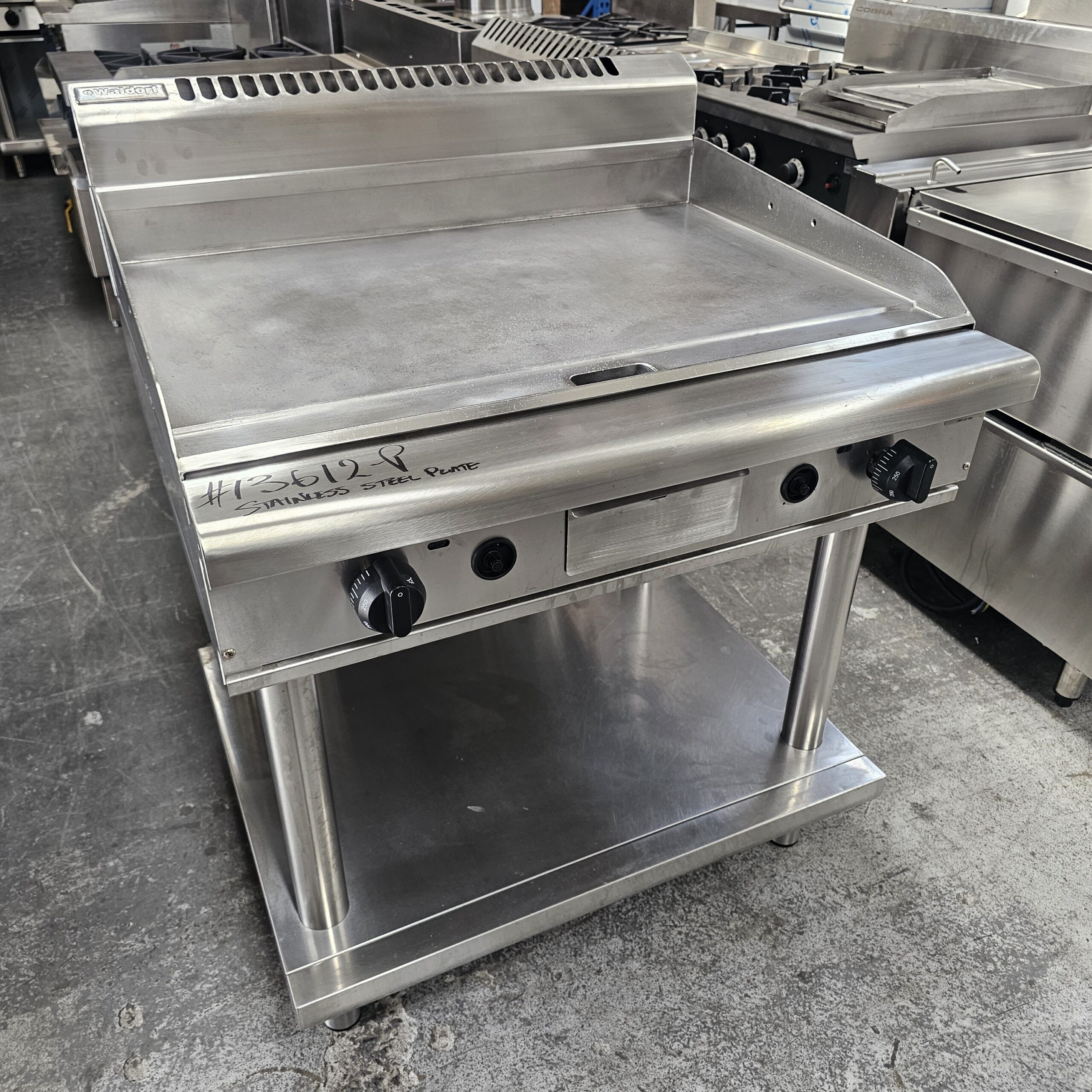 A large stainless steel commercial flat-top griddle with two control knobs, a shelf underneath, and a backsplash, situated in an industrial kitchen setting. MRCE
