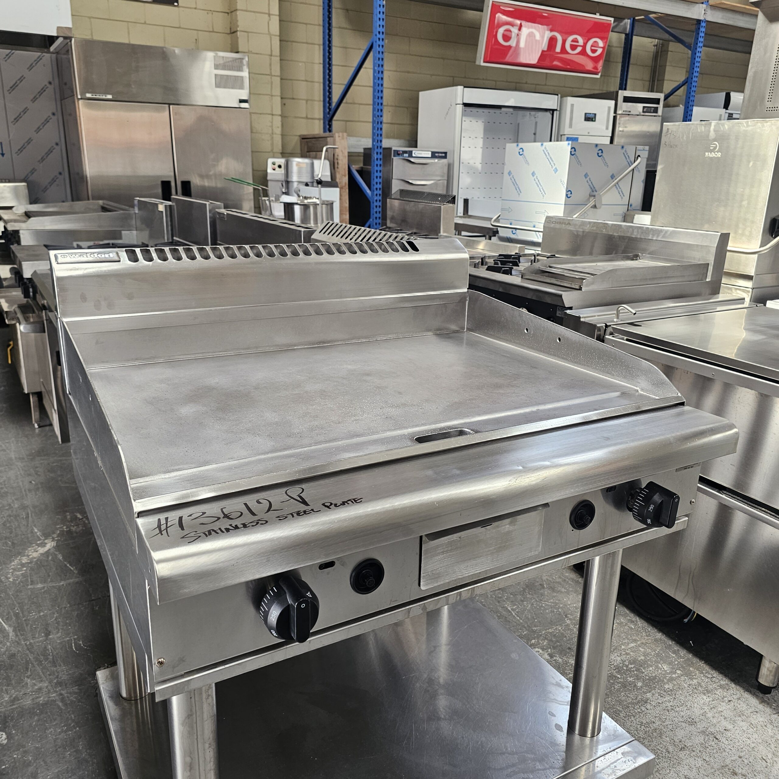 A commercial stainless steel flat-top griddle with two control knobs, labeled #35618, is displayed in a warehouse surrounded by other kitchen equipment and appliances. MRCE