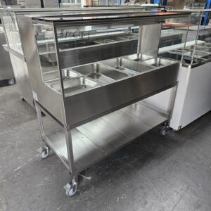 A stainless steel food service cart with a glass sneeze guard, six metal food compartments on top, and a lower storage shelf, standing on wheels in an industrial setting. MRCE