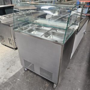A stainless steel commercial refrigerated display case with a glass top and multiple metal food trays, situated on wheels in an industrial setting. MRCE