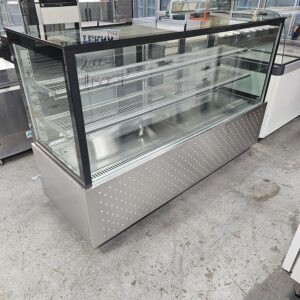 A large, empty, stainless steel and glass display case with three interior shelves stands on a concrete floor in a commercial setting. Handwritten numbers are visible on the glass front. MRCE