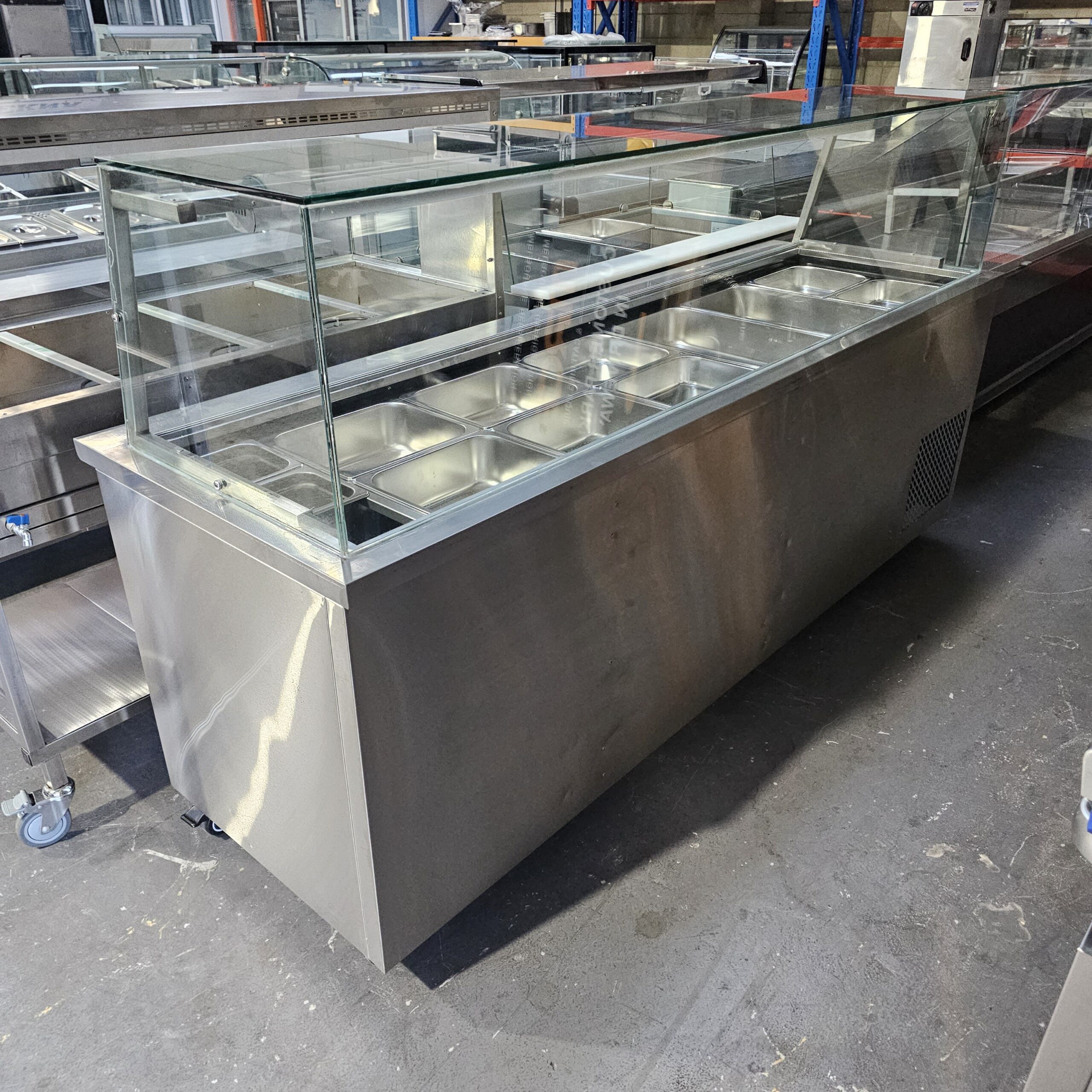 A stainless steel commercial food display counter with a glass sneeze guard and multiple empty metal food trays, situated in an industrial warehouse setting. MRCE