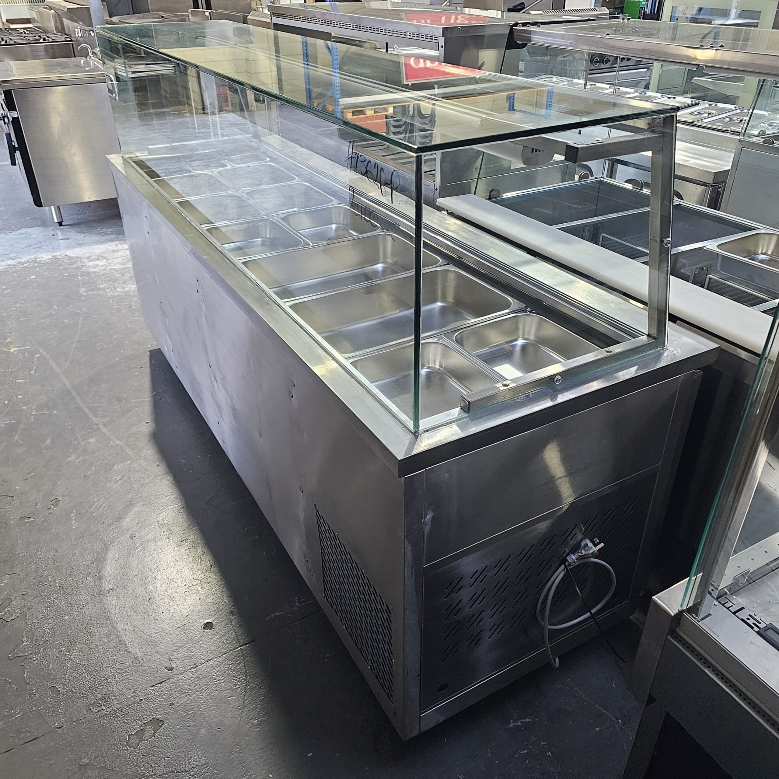 A stainless steel refrigerated food display counter with a glass sneeze guard, featuring multiple empty food trays, situated in a commercial kitchen or showroom. MRCE
