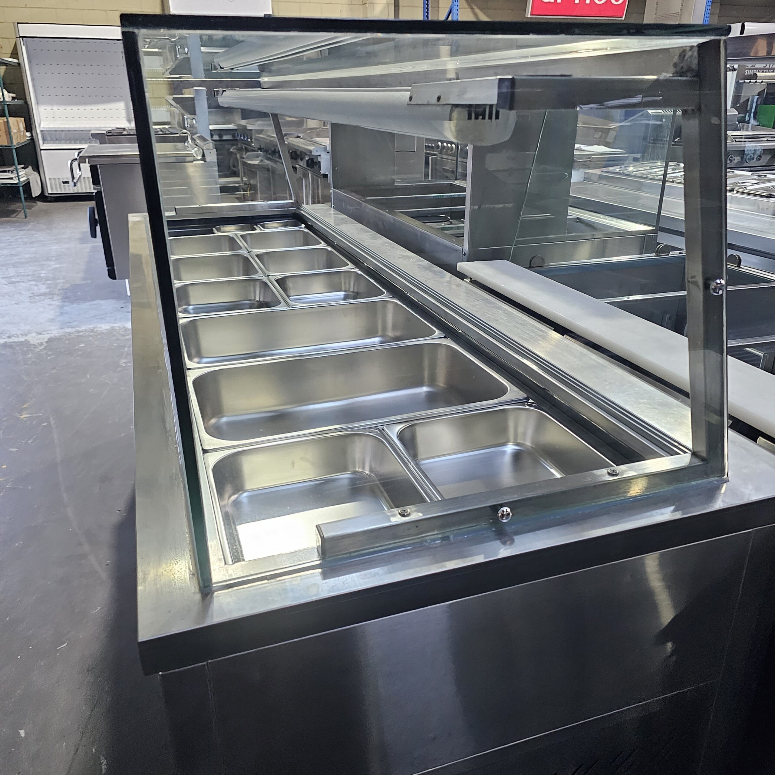 A commercial stainless steel food serving counter with an empty glass sneeze guard and multiple empty food trays, set in an industrial kitchen or warehouse environment. MRCE