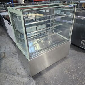 A large, empty stainless steel and glass display case with multiple shelves, standing on a concrete floor in an industrial setting surrounded by other equipment. MRCE