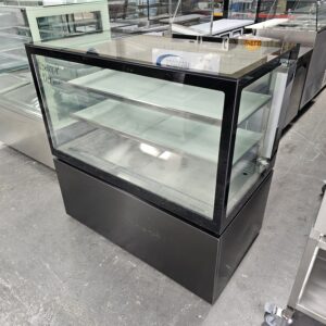 A glass display case with black framing and two interior shelves, standing on a gray concrete floor in a showroom filled with other commercial kitchen equipment. MRCE