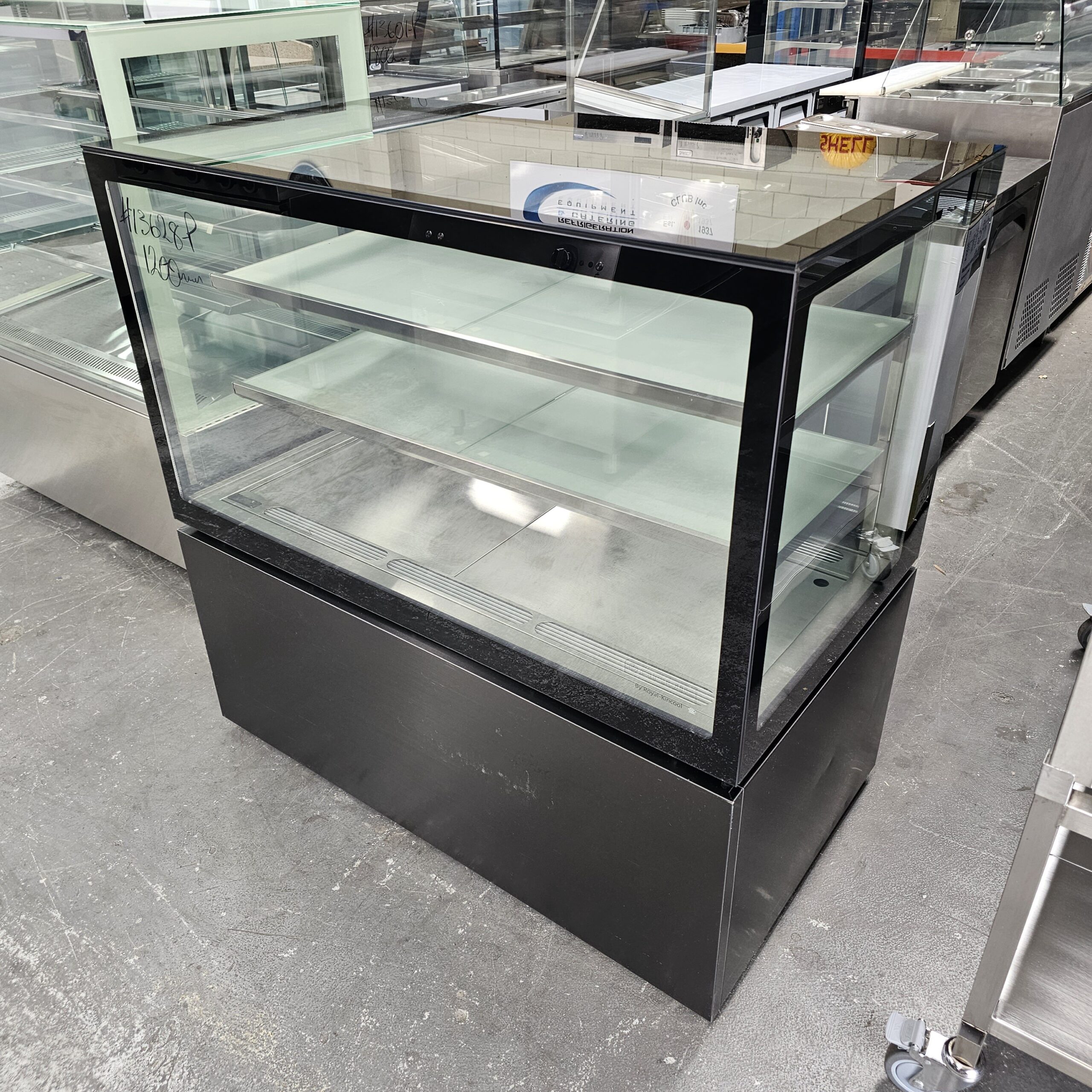 A glass display case with black framing and two interior shelves, standing on a gray concrete floor in a showroom filled with other commercial kitchen equipment. MRCE