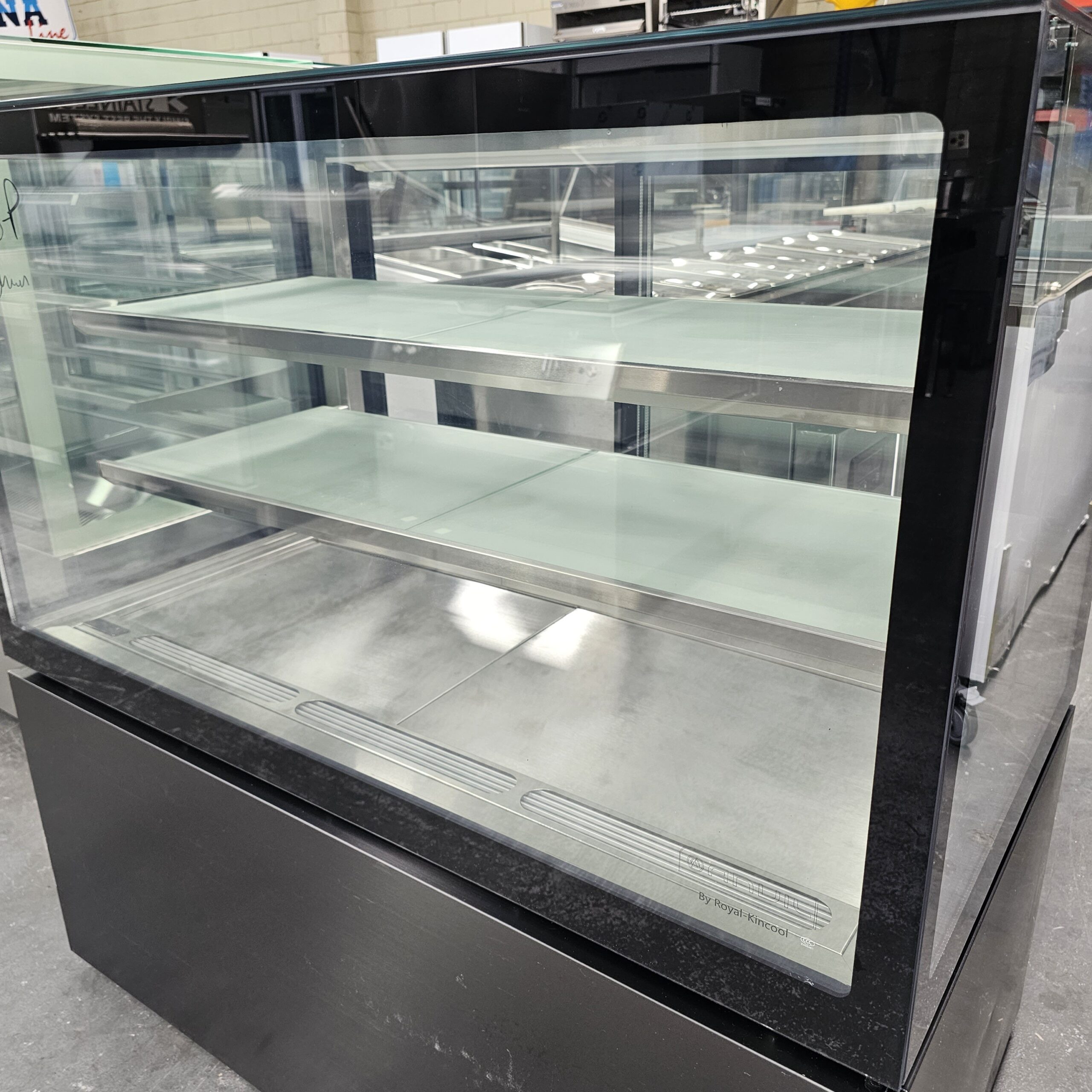 A large, empty glass display case with three shelves and a black frame is situated indoors. The case appears clean and ready for use, likely intended for displaying food items in a store or bakery. MRCE