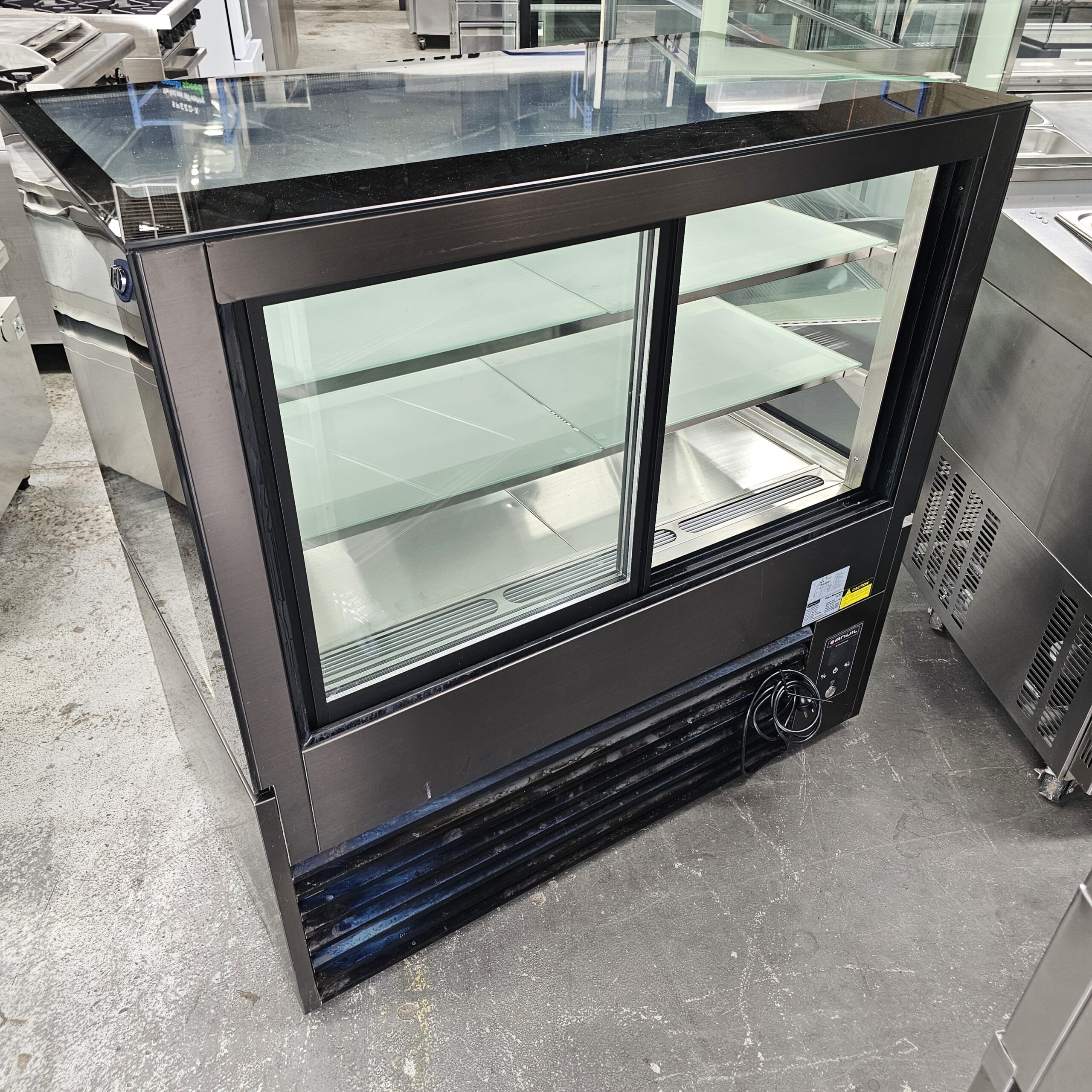 A commercial glass display refrigerator with sliding doors, black trim, and empty shelves, sitting on a concrete floor in an industrial or warehouse setting. MRCE