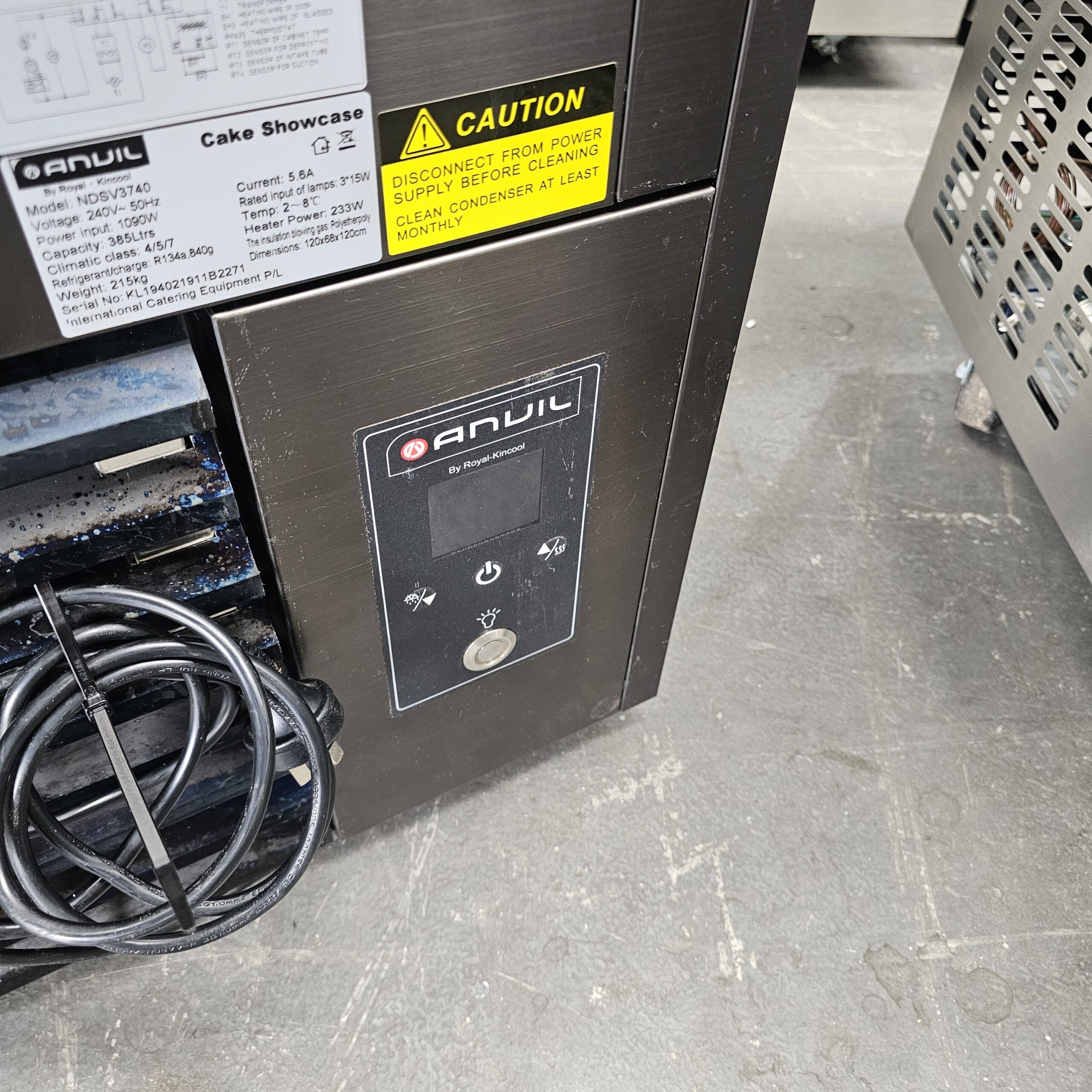 Close-up of an Anvil cake showcase control panel with caution labels and power instructions. A coiled black cable and metal surfaces are also visible on a gray concrete floor. MRCE