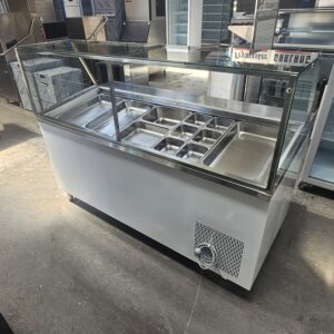A commercial stainless steel display freezer with a glass top and multiple food compartments is shown in a store or warehouse setting, standing on a concrete floor. MRCE