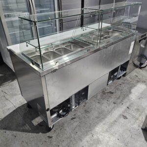 A stainless steel commercial food prep station with a glass sneeze guard, several metal food pans, and a refrigerated base, placed on a concrete floor in an industrial kitchen setting. MRCE