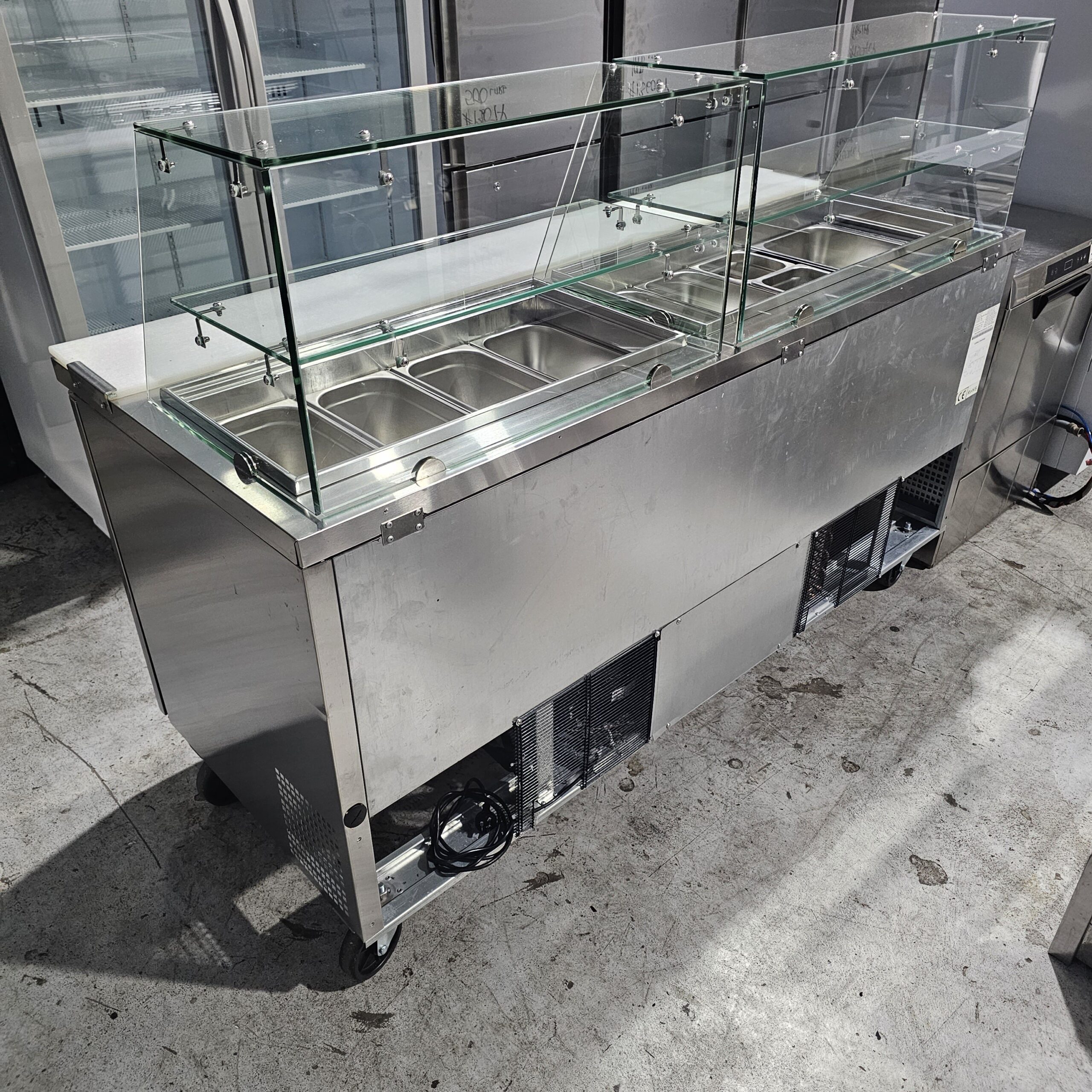 A stainless steel commercial food prep station with a glass sneeze guard, several metal food pans, and a refrigerated base, placed on a concrete floor in an industrial kitchen setting. MRCE