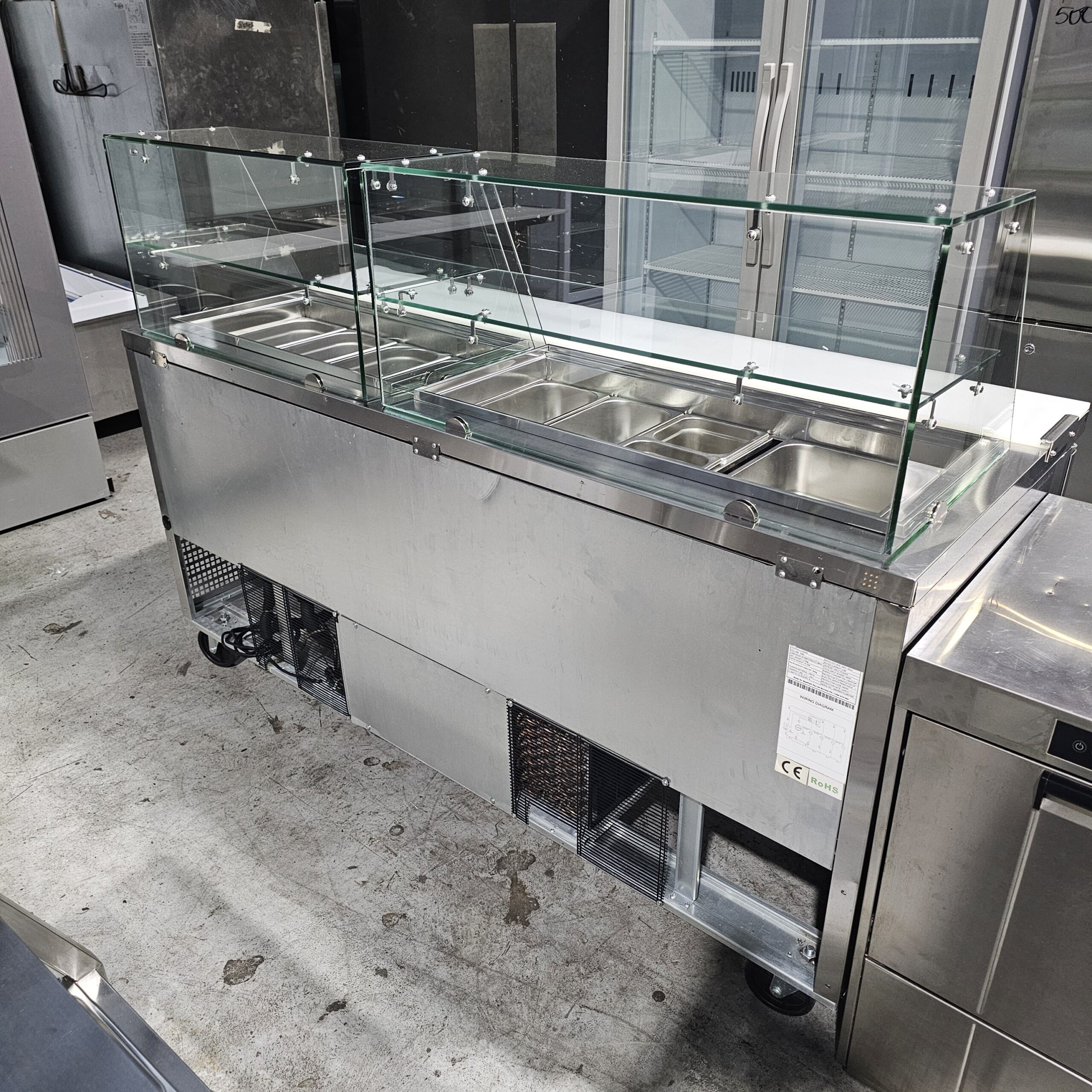 A stainless steel commercial food service counter with a glass sneeze guard, featuring multiple food trays for hot or cold items, set on a concrete floor in a kitchen or industrial setting. MRCE