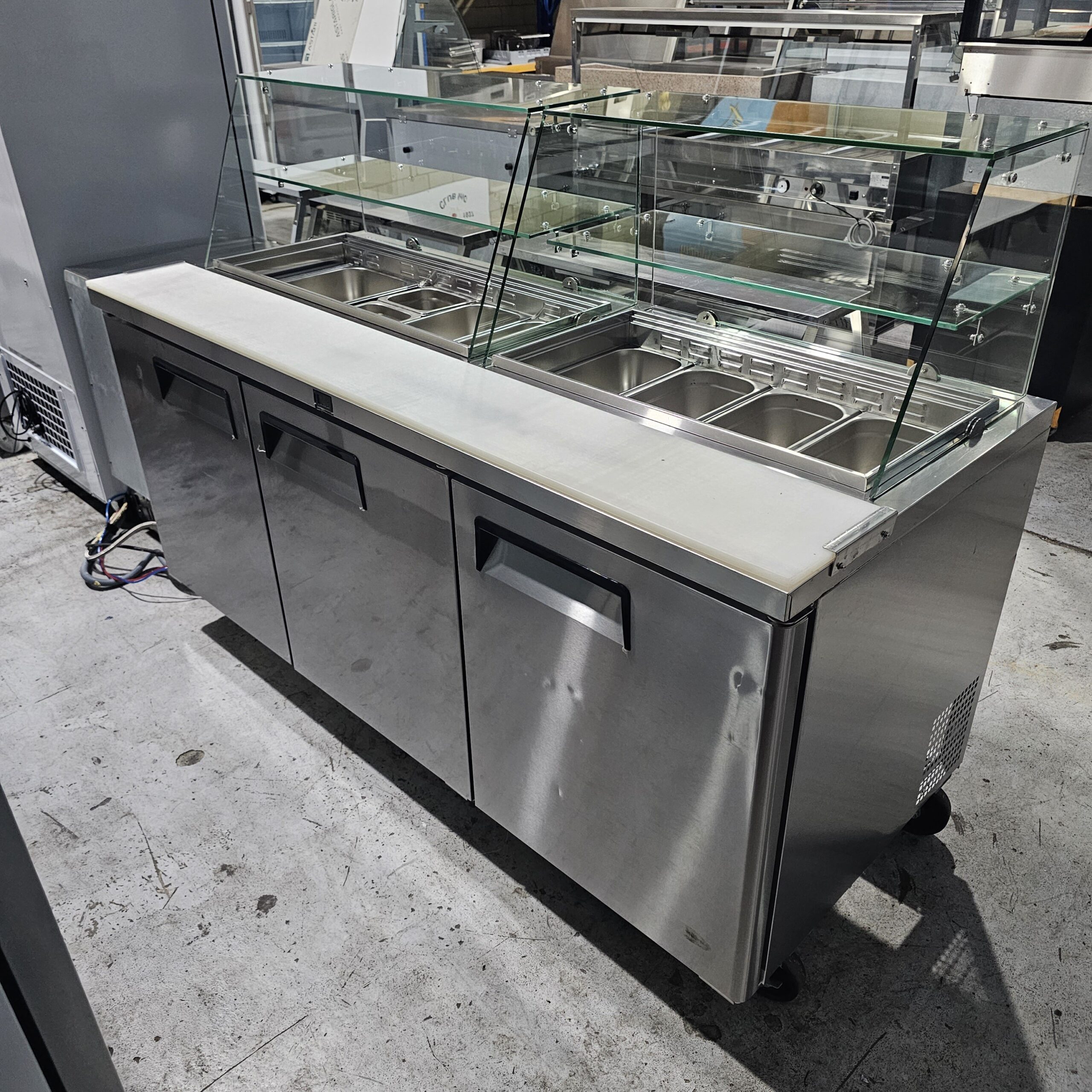 A stainless steel commercial food prep station with three lower storage compartments, a white countertop, and a glass sneeze guard covering six empty food pans on top. The unit is on wheels and set in an industrial space. MRCE