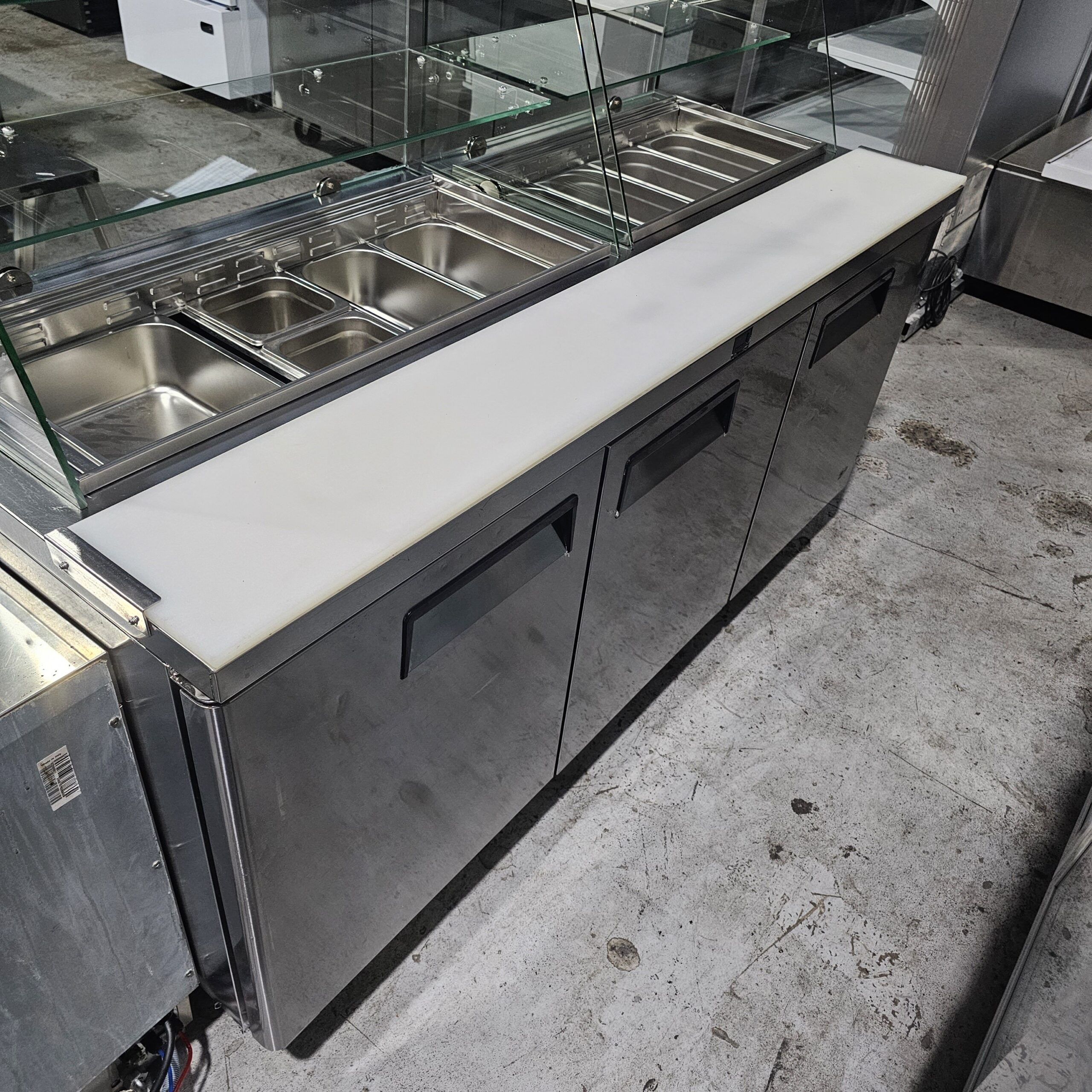 A stainless steel commercial kitchen prep station with a white cutting board, refrigerated drawers below, and multiple metal food pans under a glass shield, set on a concrete floor. MRCE