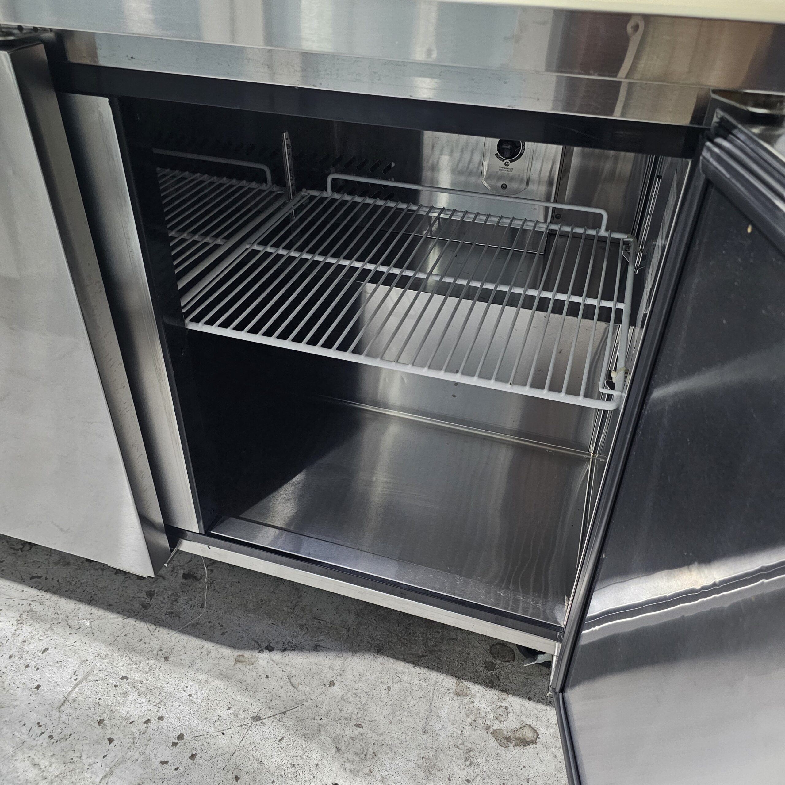 A stainless steel commercial refrigerator with its double doors open, showing empty wire shelves inside and a clean interior. The appliance is situated on a concrete floor. MRCE