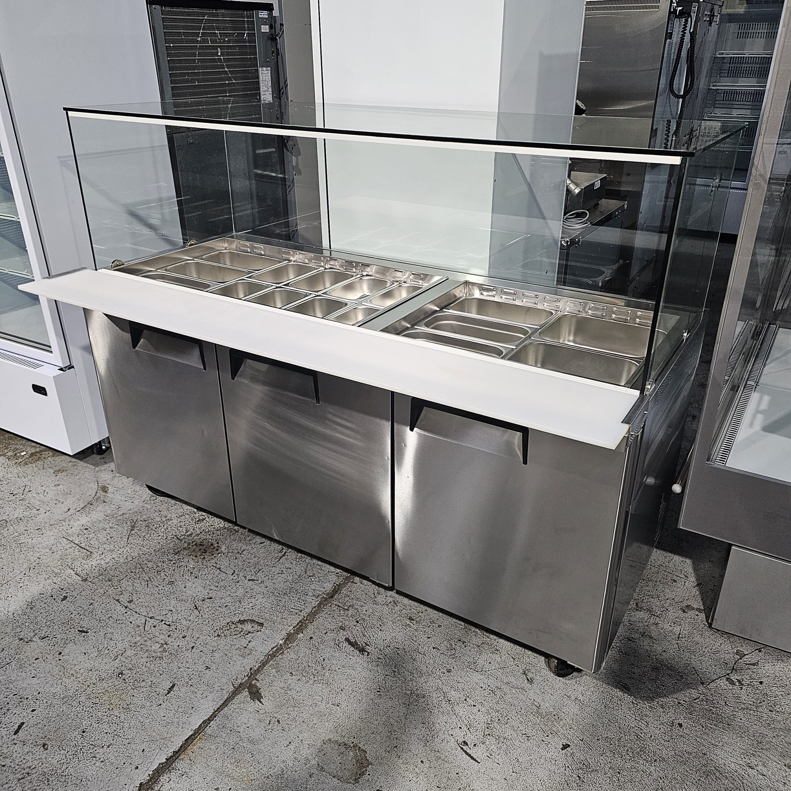 A stainless steel food service counter with a glass sneeze guard and multiple metal food trays, designed for serving hot or cold food in a commercial setting. MRCE