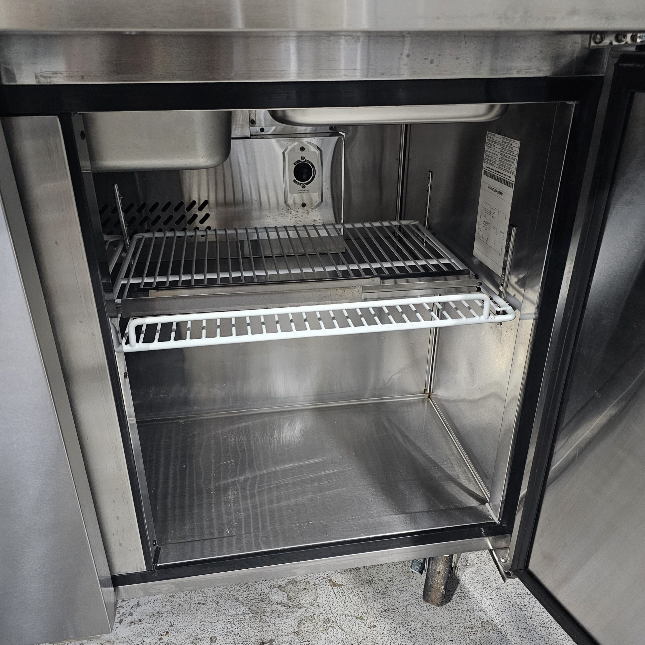 Open stainless steel commercial refrigerator with a single wire shelf inside, empty interior, and open door revealing a clean, metallic surface and ventilation grates at the back. MRCE