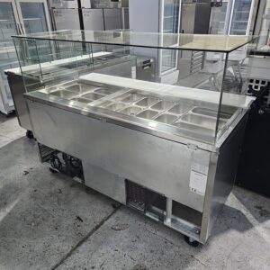 A stainless steel commercial food display counter with a glass sneeze guard and multiple metal food trays, situated in an industrial kitchen or showroom setting. MRCE