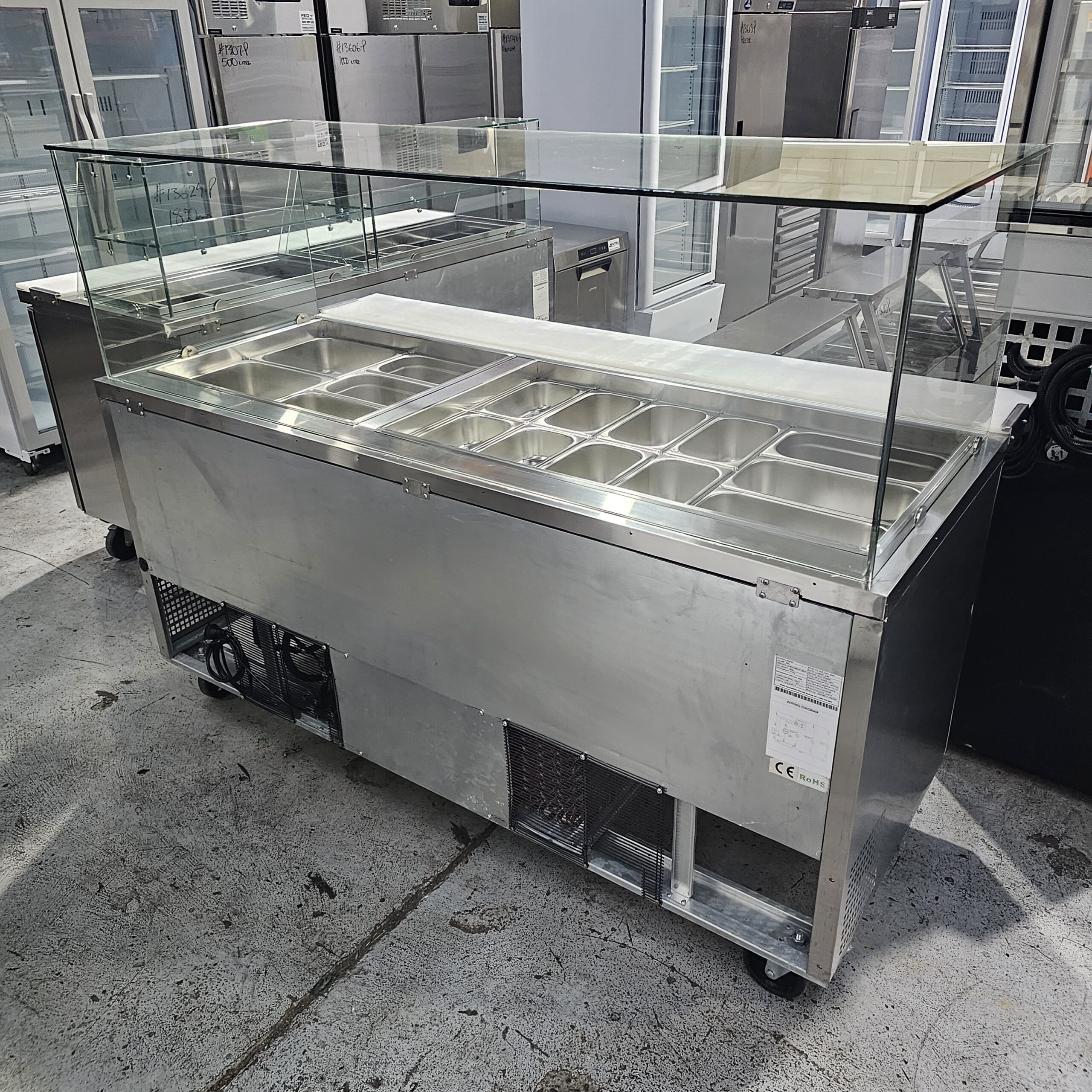 A stainless steel commercial food display counter with a glass sneeze guard and multiple metal food trays, situated in an industrial kitchen or showroom setting. MRCE