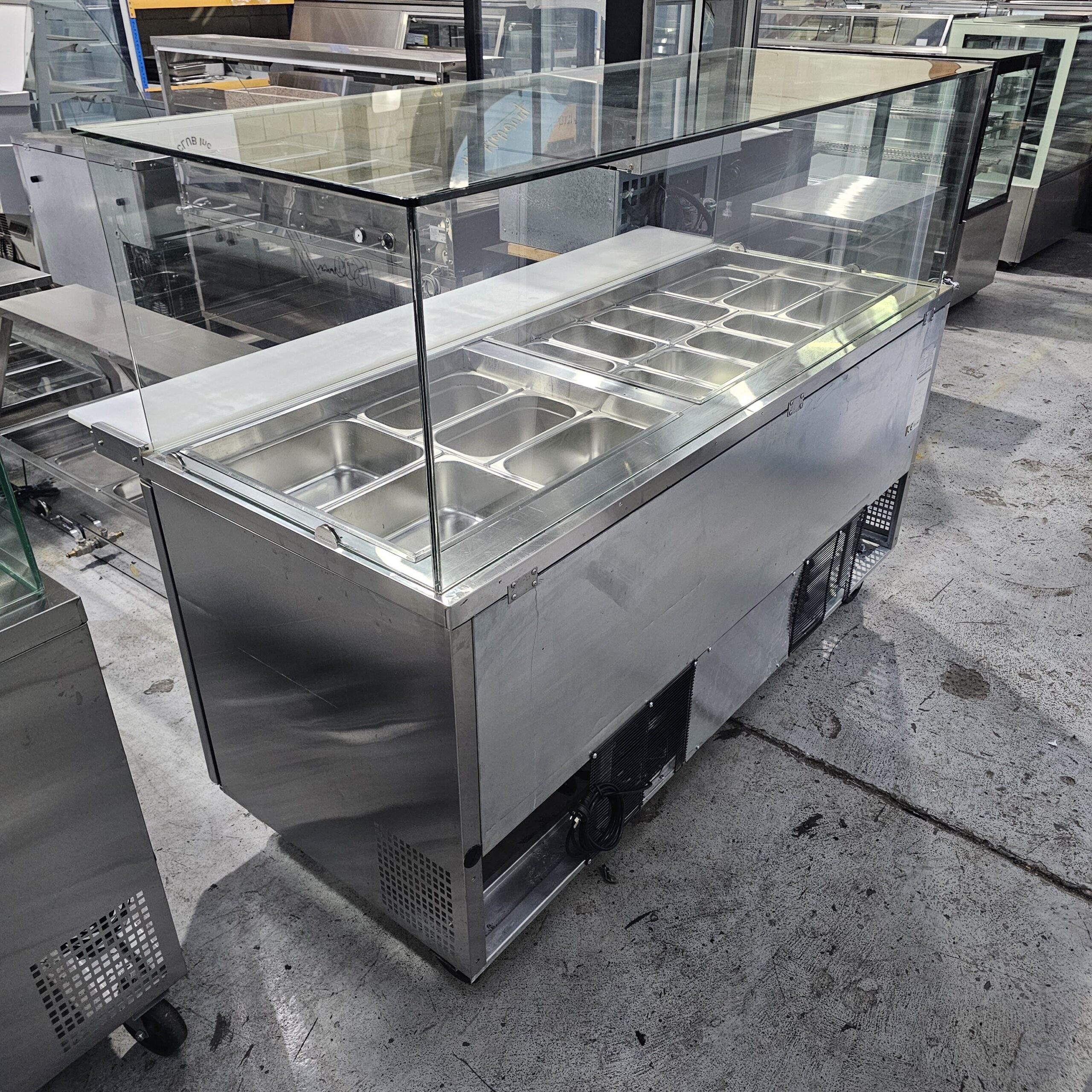 A commercial stainless steel food display counter with a glass cover, featuring multiple metal food pans, situated in an industrial warehouse setting. MRCE