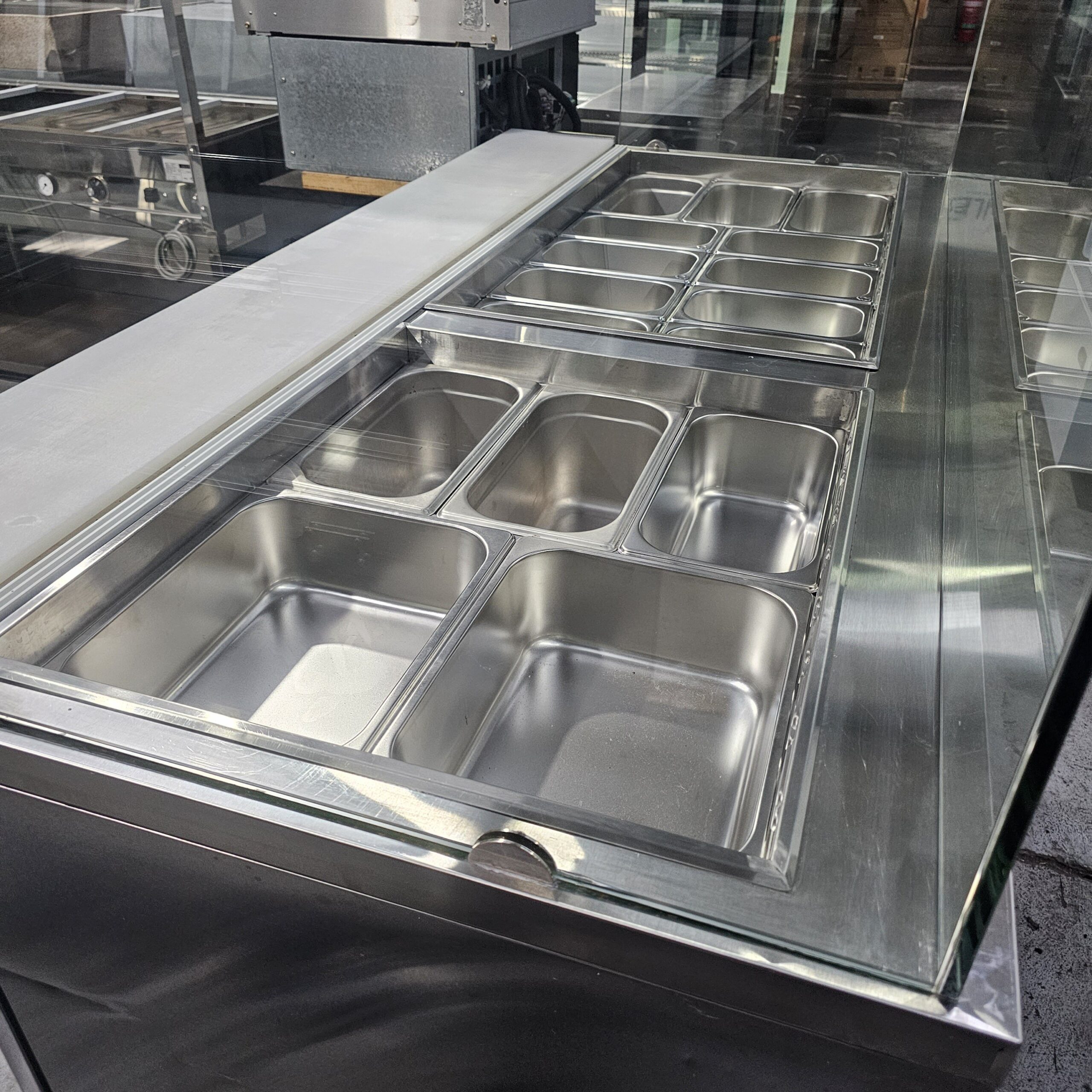 A stainless steel food service counter with several empty metal food trays, covered by a glass sneeze guard. The trays are clean and arranged neatly, and the counter is in a commercial kitchen or cafeteria setting. MRCE