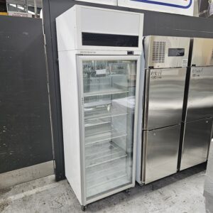 A tall, white commercial display refrigerator with a glass door and empty metal shelves stands next to stainless steel appliances in a store or warehouse setting. MRCE