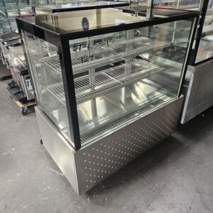 A stainless steel and glass refrigerated display case with three shelves, situated in a warehouse. The case has a perforated lower front panel and clear glass sides and top. MRCE