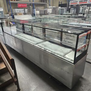 A large commercial refrigerated display case with glass panels sits in a warehouse, surrounded by other commercial kitchen equipment. Some items are inside the case, and a handwritten sign is visible on the glass. MRCE