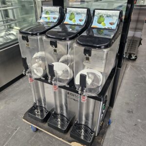 A commercial beverage dispenser with three empty clear containers and black lids sits on a pallet in an industrial kitchen setting. Each container has a spout and colorful fruit graphics on top. MRCE