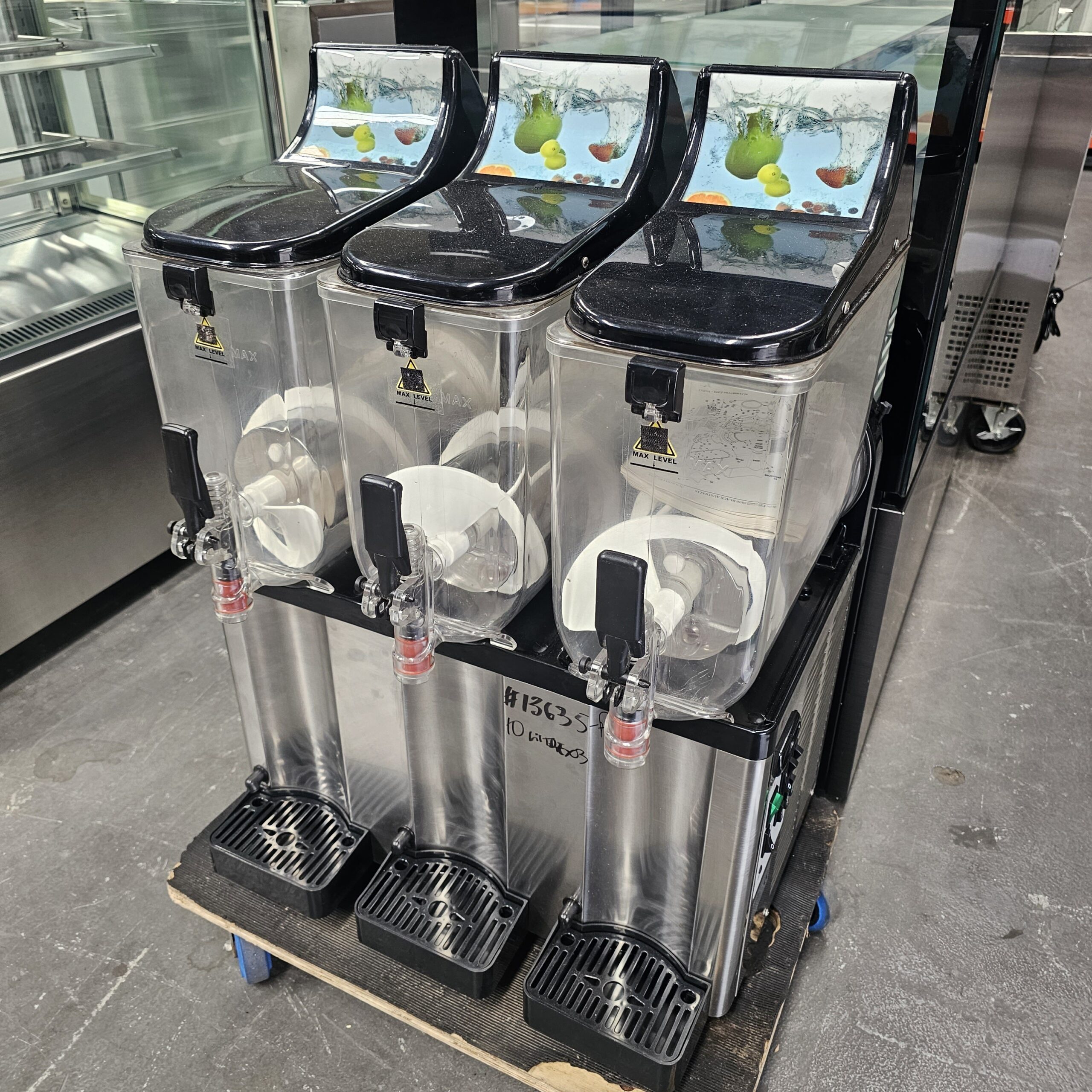 A commercial beverage dispenser with three empty clear containers and black lids sits on a pallet in an industrial kitchen setting. Each container has a spout and colorful fruit graphics on top. MRCE