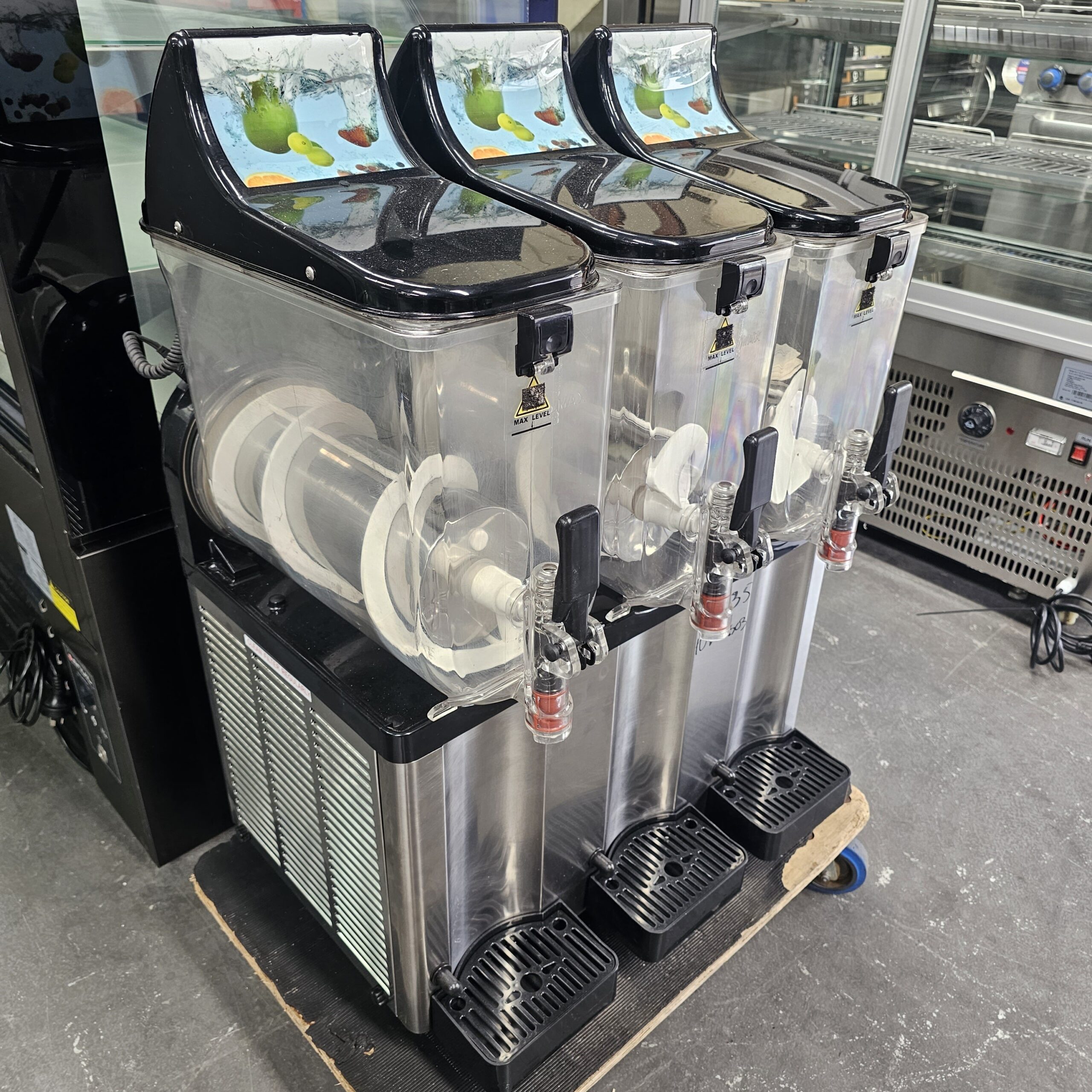 Three side-by-side commercial slush machines with clear tanks and black lids, each with a spout for dispensing frozen drinks, are placed on a black base in a store or cafe setting. MRCE