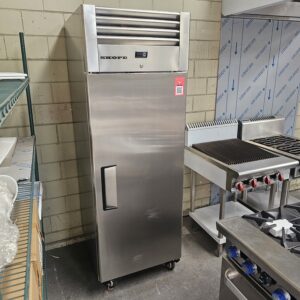 A large, stainless steel commercial refrigerator with the brand name SKOPE stands in a kitchen near a metal shelf and cooking appliances. It has a single door and is on wheels, set against a tiled wall. MRCE