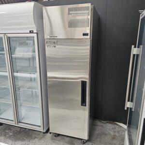 A tall, stainless steel, single-door commercial refrigerator stands between other appliances in a store. A price ($1359.99) and 650 Litre are written in black marker on the door. The refrigerator is on small wheels. MRCE