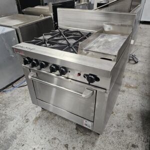 A commercial stainless steel gas range with four burners, a griddle, and an oven below, situated in a professional kitchen setting with other appliances around. MRCE