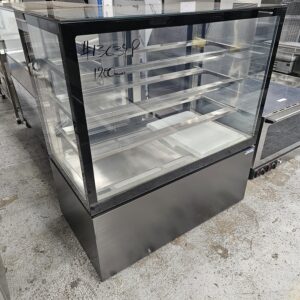 A glass display case with three shelves and a stainless steel base, standing on a concrete floor in what appears to be a commercial kitchen or showroom. Handwritten text is visible on the glass. MRCE