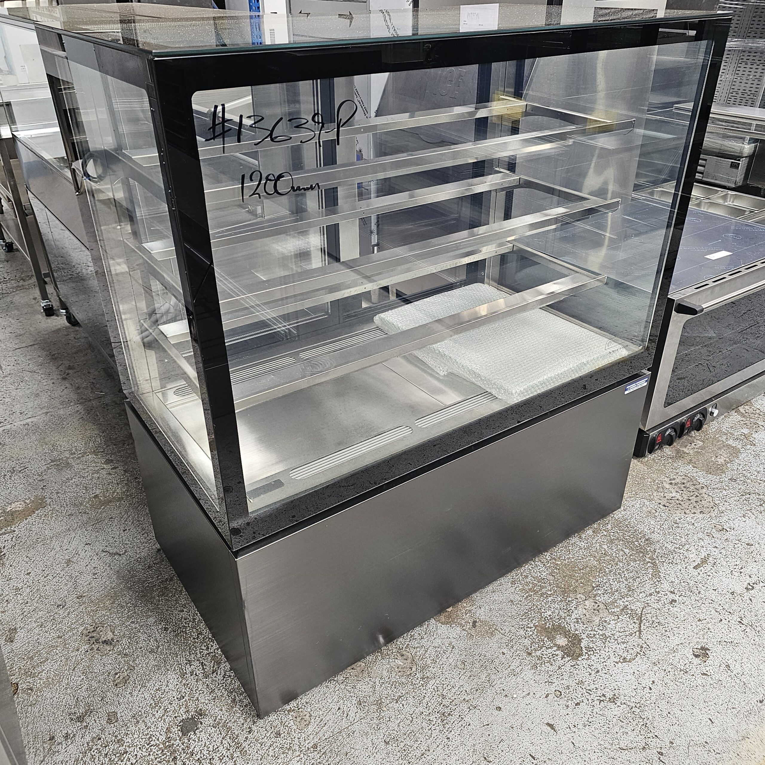 A glass display case with three shelves and a stainless steel base, standing on a concrete floor in what appears to be a commercial kitchen or showroom. Handwritten text is visible on the glass. MRCE