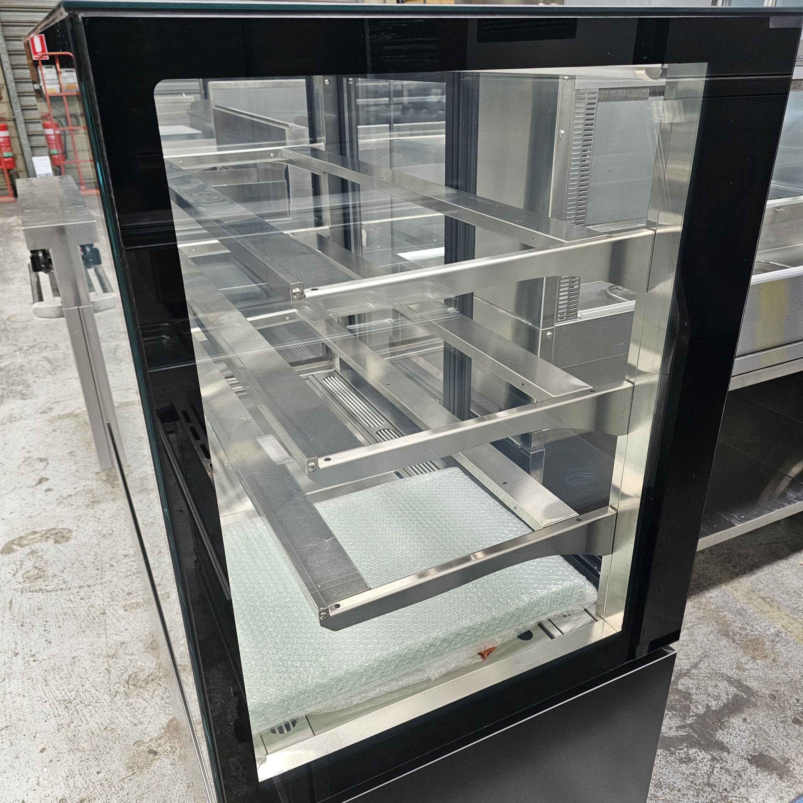 A glass display case with multiple empty metal shelves is shown in an indoor setting with a concrete floor. The case appears new and unused, with bubble wrap on the bottom shelf. MRCE