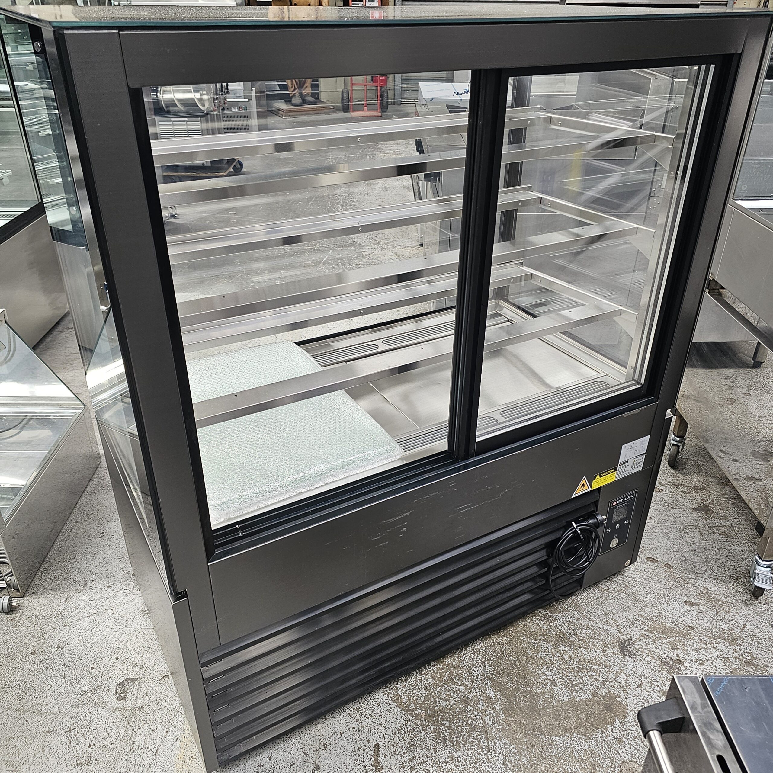 A black refrigerated display case with glass panels and several shelves stands on a concrete floor, designed for showcasing food or beverages in a commercial setting. MRCE