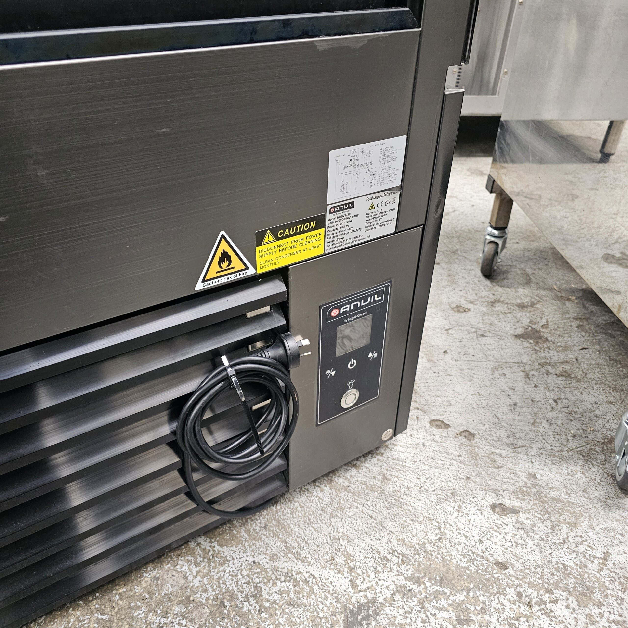 A close-up of a commercial refrigeration unit on a concrete floor, featuring warning labels, a digital control panel with the brand SANUL, and a coiled black power cord hanging on the front. MRCE