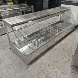 A large, empty, stainless steel and glass display case sits on a concrete floor in a commercial setting, likely used for showcasing food items. The case has sliding doors and labeled writing on the glass. MRCE