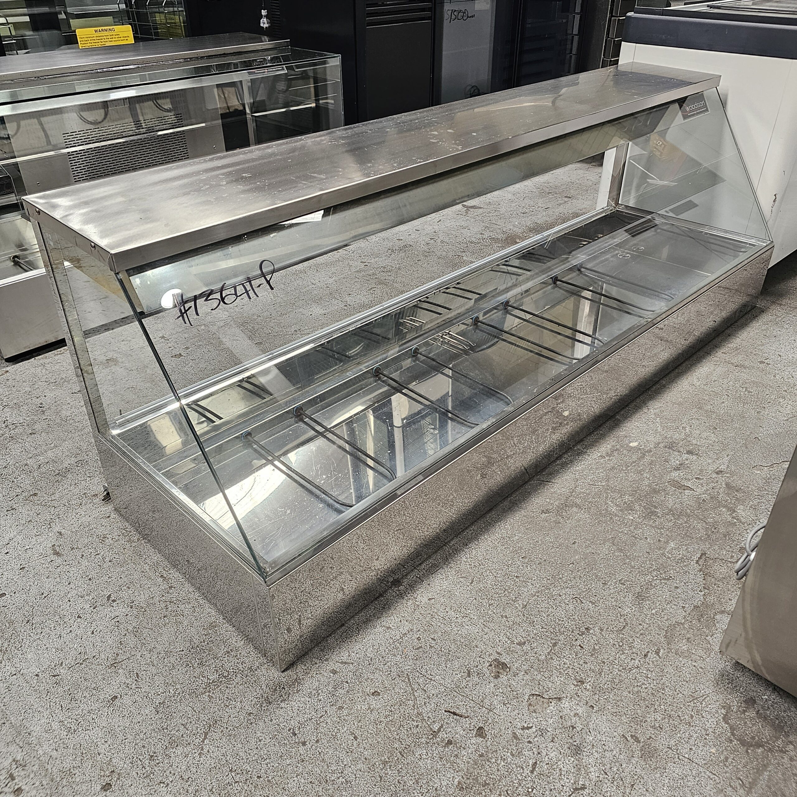 A large, empty, stainless steel and glass display case sits on a concrete floor in a commercial setting, likely used for showcasing food items. The case has sliding doors and labeled writing on the glass. MRCE