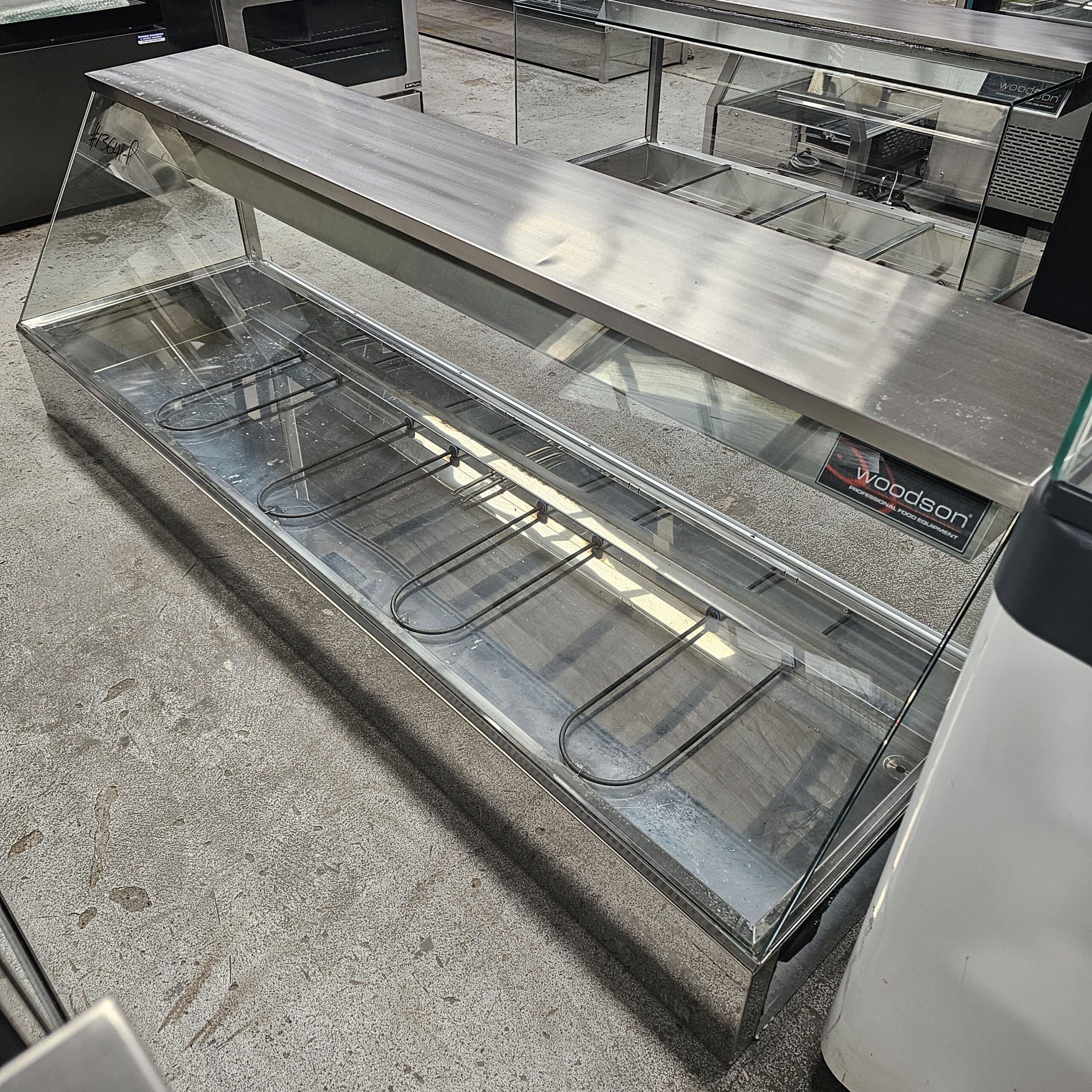 A large empty stainless steel and glass food display case with heating elements on the bottom, placed on a concrete floor in what appears to be a commercial or industrial setting. MRCE