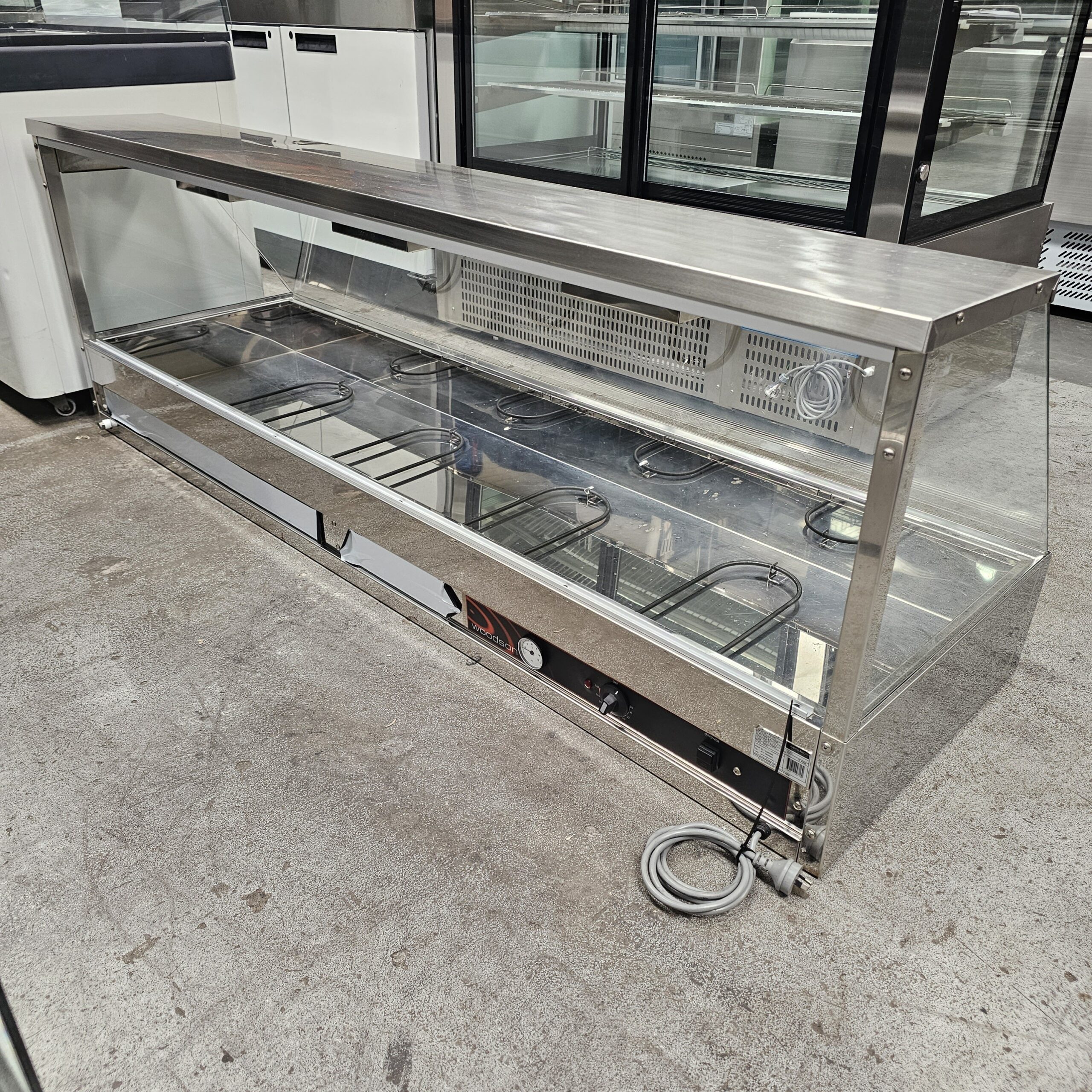 A large, empty stainless steel and glass display case with electrical cords sits on a concrete floor in a commercial setting. MRCE