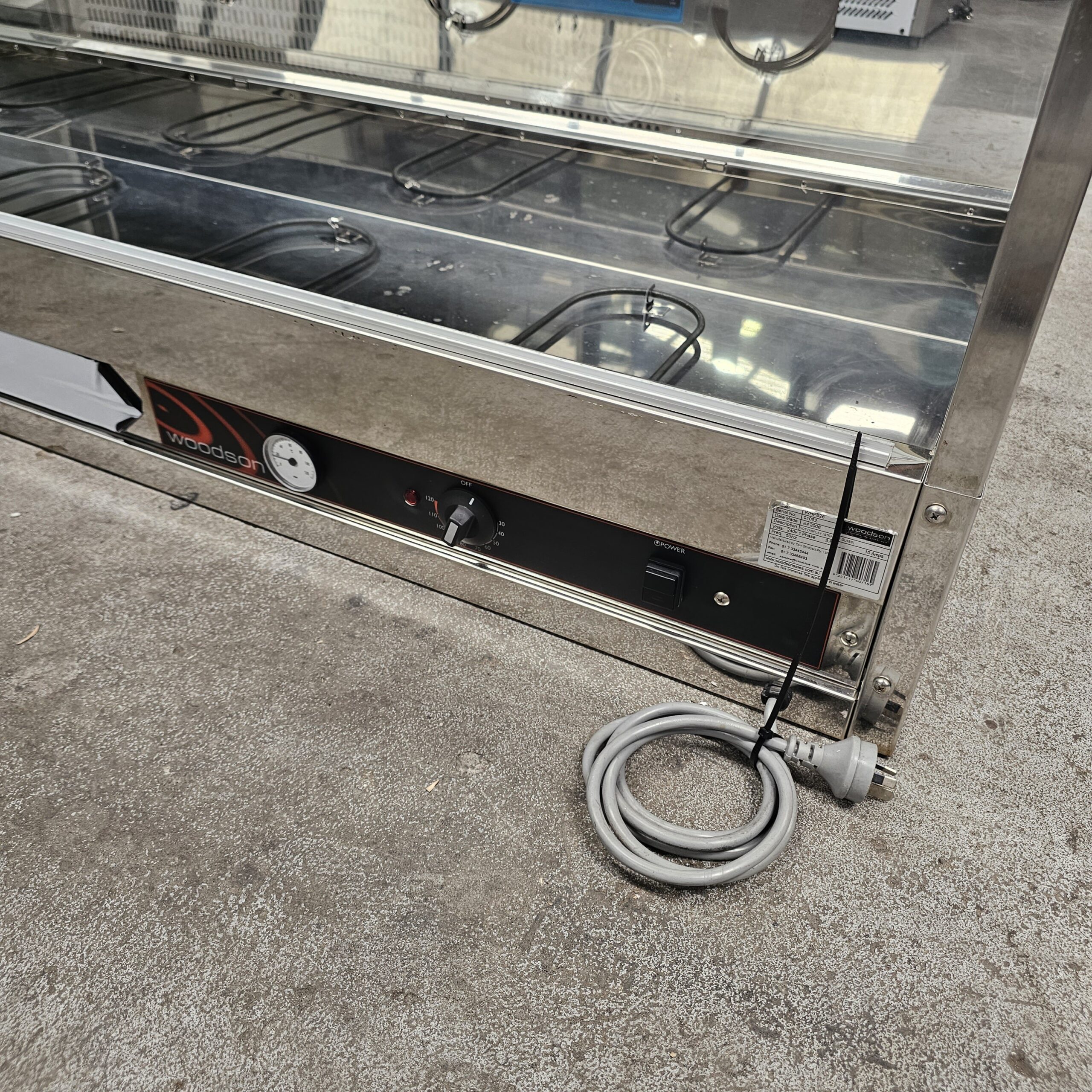 A stainless steel commercial warming display unit sits on a concrete floor. The control panel features a dial, a gauge, and a power cord coiled neatly in front. MRCE