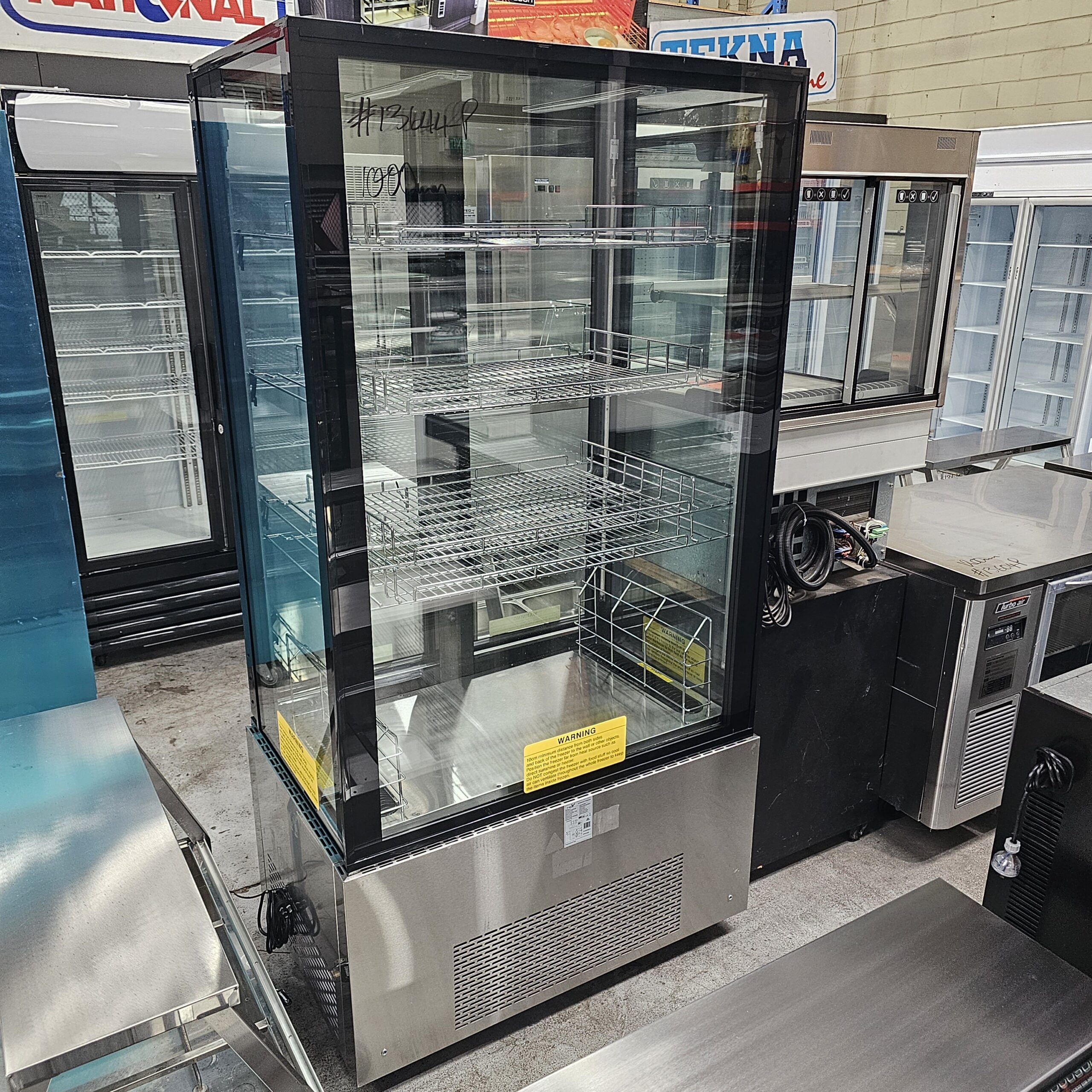 A commercial glass display refrigerator with multiple wire shelves stands empty in a showroom with other refrigeration units and stainless steel appliances around it. MRCE