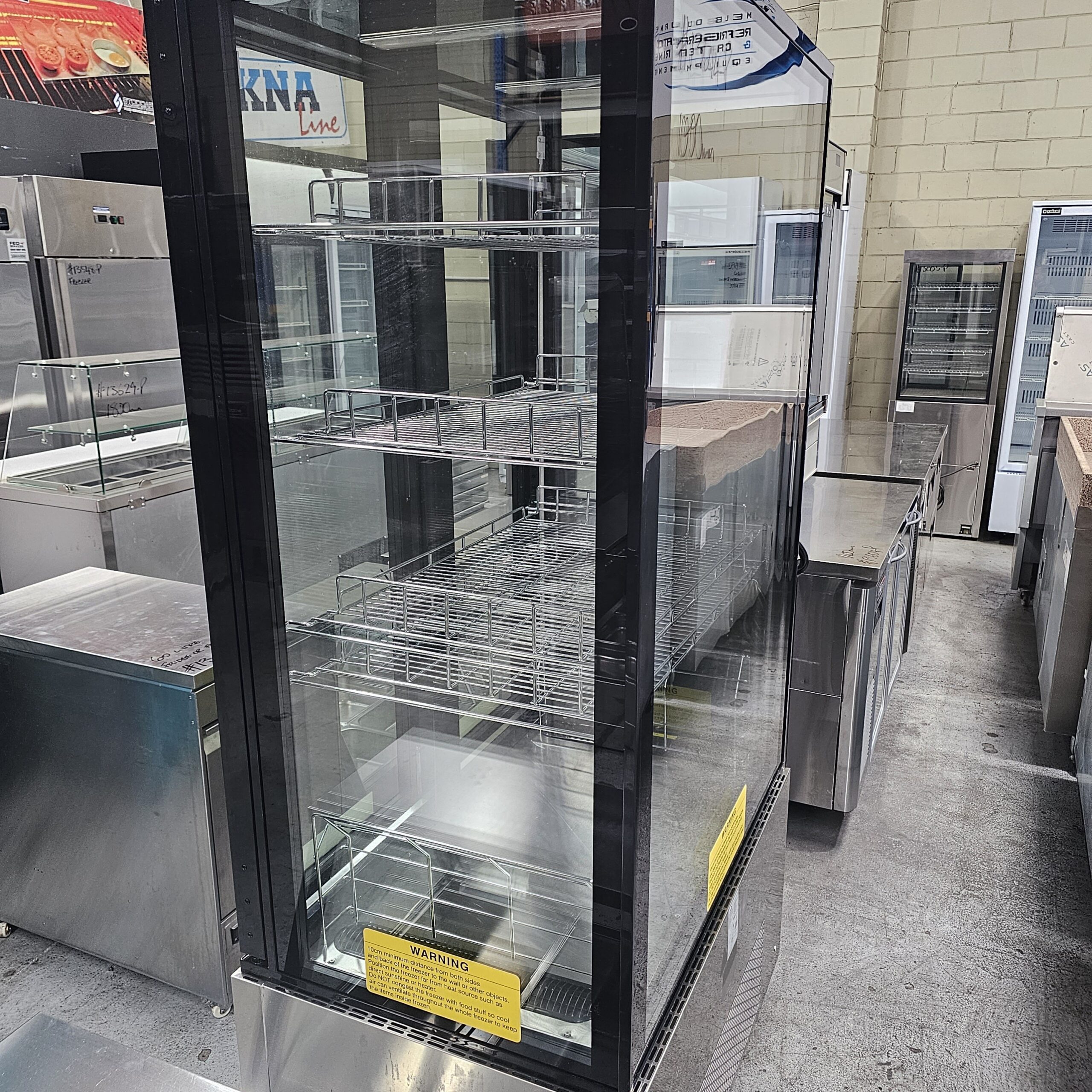 A large, empty glass display refrigerator with wire shelves stands in a commercial kitchen or equipment showroom. Stainless steel counters and appliances surround it, and a yellow warning label is visible on the front. MRCE