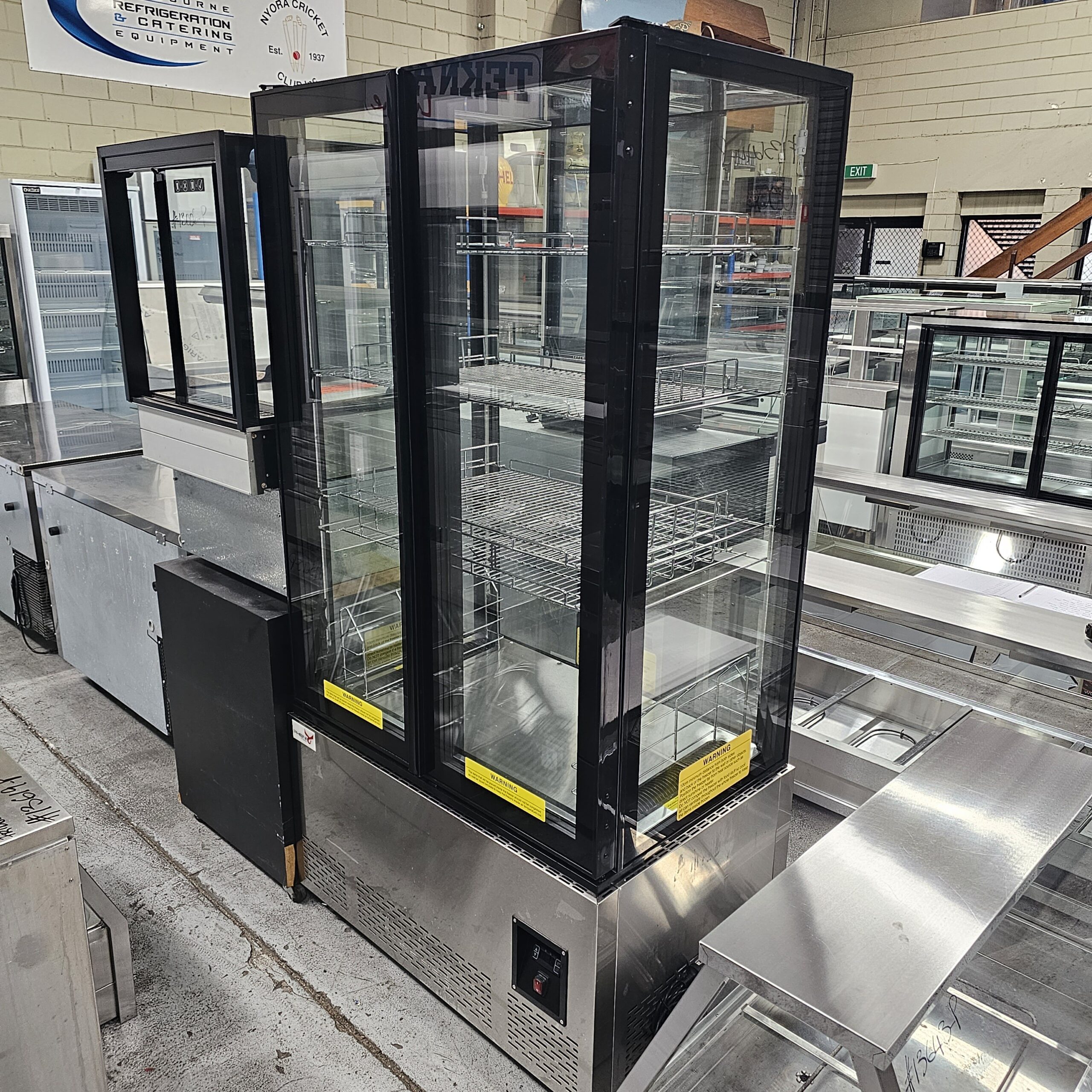 A glass-door refrigerated display case with multiple shelves stands in a warehouse among other commercial kitchen equipment and stainless steel counters. MRCE